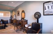 Small salon with several vintage styling stations, chairs, mirrors, and a hooded hair dryer along a light-blue wall.