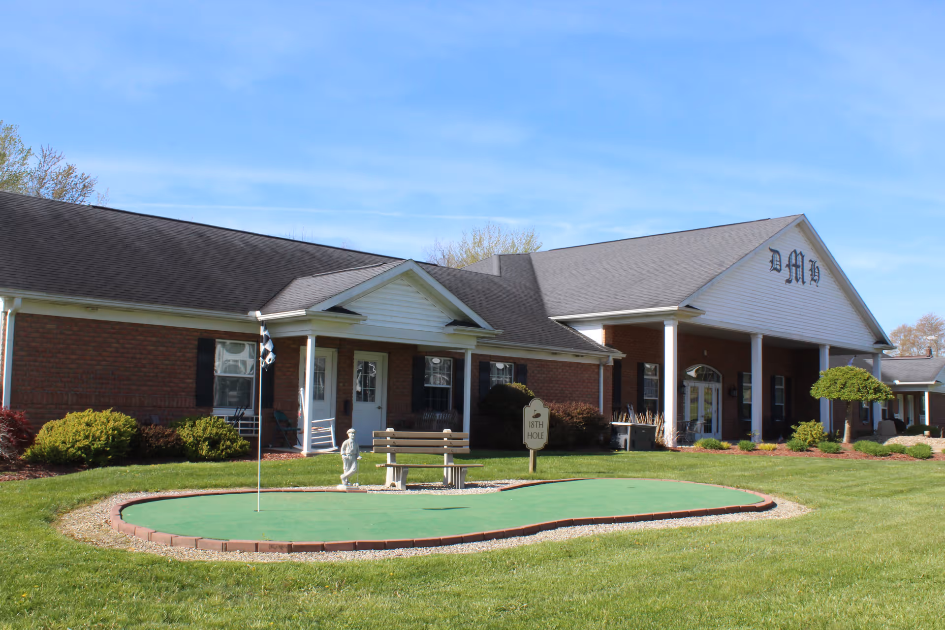 Exterior view of a single-story brick building with white columns and a black roof under a clear blue sky. In front of the building is a small artificial putting green with a flag, a bench, and a small statue. The building has the letters 'DMH' on the front gable and is surrounded by well-maintained grass and shrubs.