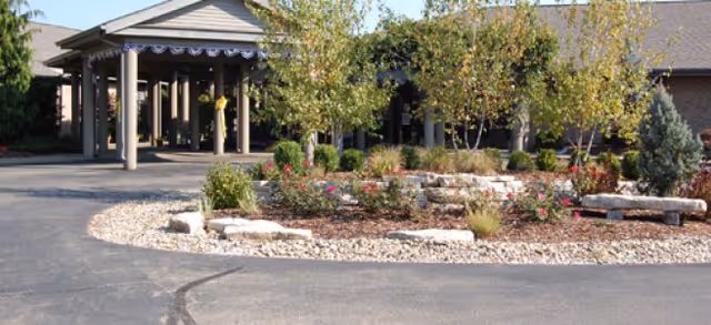 Entrance area of a senior living facility with a covered drop-off zone supported by columns, surrounded by landscaped garden beds with small trees, shrubs, and flowers, and a paved driveway.