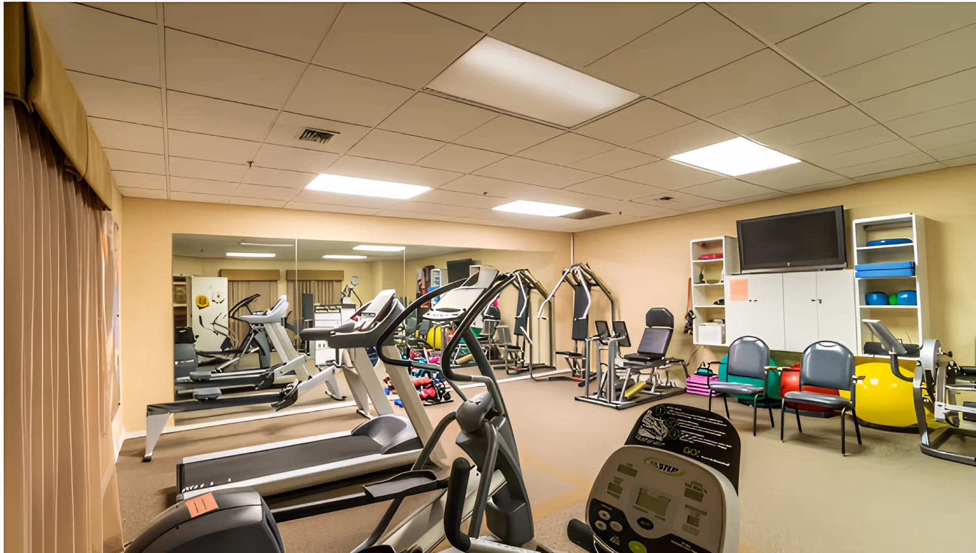 Interior view of a fitness room in a senior living facility with exercise equipment including treadmills, elliptical machines, weight machines, and exercise balls. There is a large mirror on one wall, shelves with fitness accessories, chairs, and a wall-mounted television.