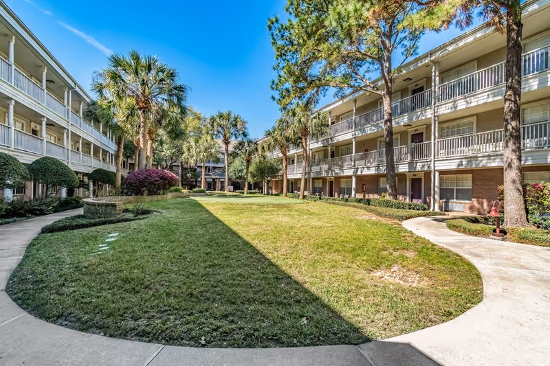 Outdoor courtyard area of a senior living facility with a well-maintained grassy lawn, palm trees, and other greenery. The courtyard is surrounded by a three-story building with balconies and walkways. The sky is clear and blue.
