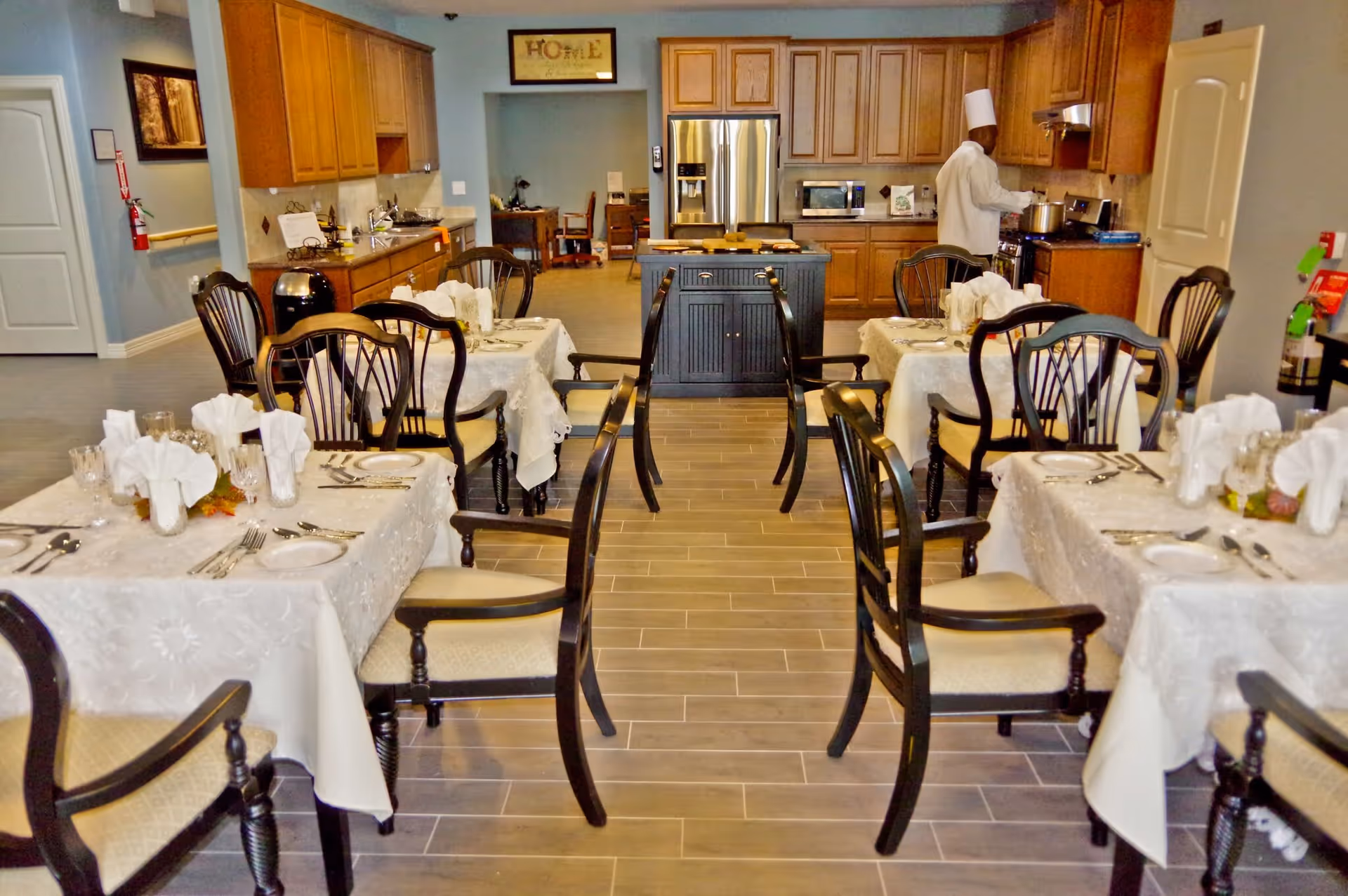 A dining area with several tables covered in white tablecloths, each set with plates, glasses, and folded napkins. Wooden chairs with cushioned seats surround the tables. In the background, a chef wearing a white uniform and hat is cooking in a kitchen area with wooden cabinets and stainless steel appliances.
