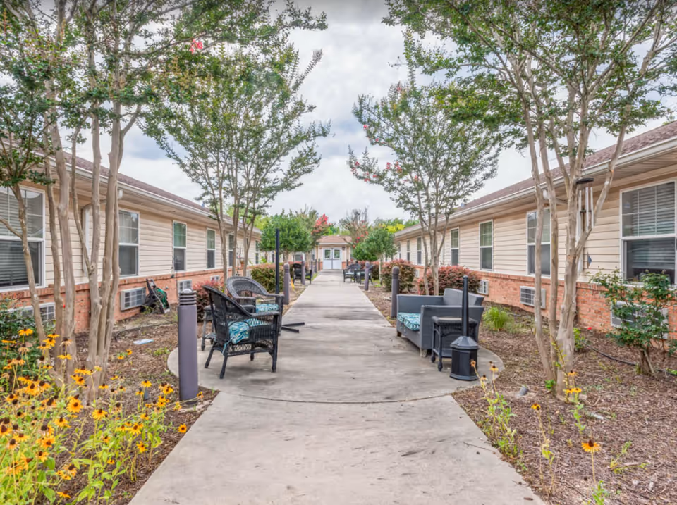 Outdoor walkway between two single-story buildings with beige siding and brick accents, lined with trees, flower beds, and outdoor seating including chairs and small tables.