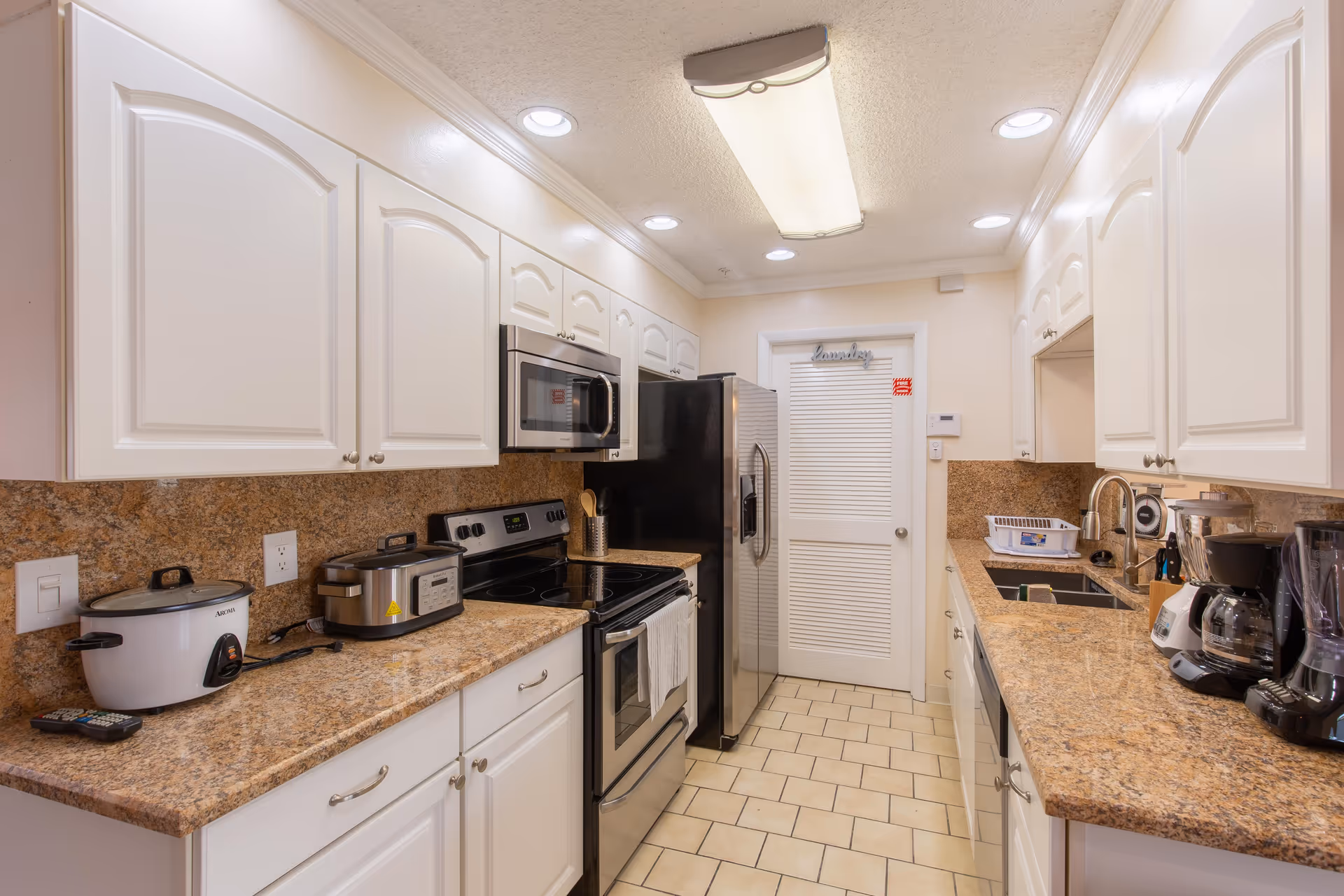 A clean and well-lit kitchen with white cabinets and granite countertops. The kitchen features a black electric stove with an oven, a stainless steel microwave above it, and a large stainless steel refrigerator. On the left countertop, there is a rice cooker and a slow cooker. On the right countertop, there is a coffee maker, a blender, a dish rack, and a double sink. The floor is tiled with beige tiles, and there is a door labeled 'laundry' at the end of the kitchen.