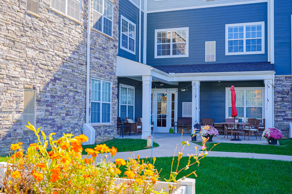 Outdoor patio area of a senior living facility with stone and blue siding exterior walls, several windows, patio tables and chairs, a red umbrella, and colorful flowers in the foreground.