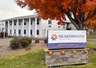 Exterior view of Hearthstone Senior Living facility with a large tree with red autumn leaves in the foreground and a sign displaying the facility name and services offered.