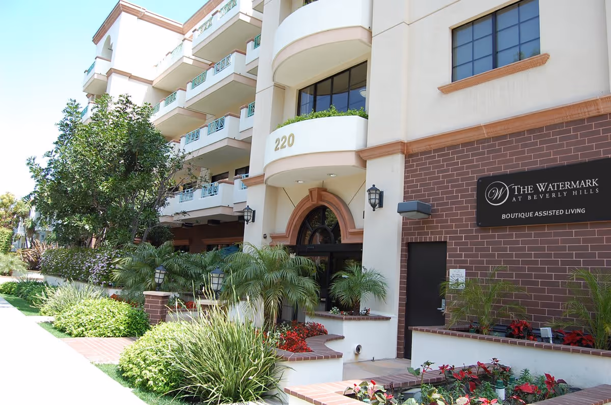 Exterior view of The Watermark at Beverly Hills assisted living facility showing the building entrance with the address number 220, landscaped garden with various plants and flowers, and a sign that reads 'The Watermark at Beverly Hills Boutique Assisted Living'.