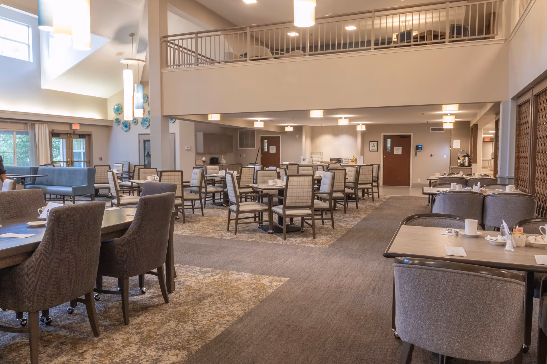 Spacious dining room in a senior living facility with multiple tables and chairs arranged neatly. The room features high ceilings, large windows allowing natural light, modern light fixtures, and a second-floor balcony overlooking the area. Tables are set with cups, napkins, and condiments.