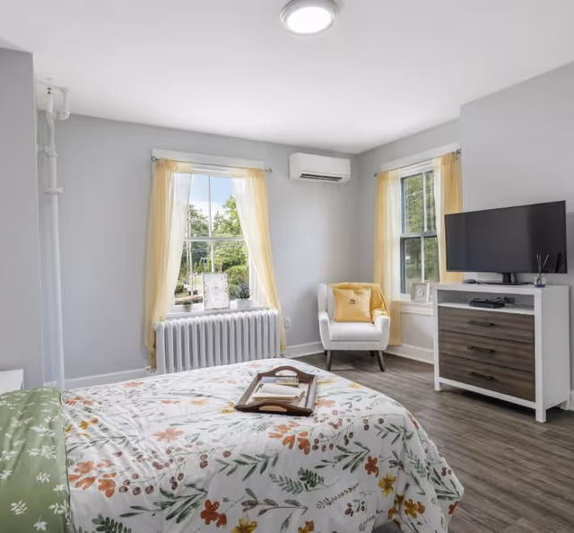 Bright, neatly staged bedroom with a floral bedspread, an armchair by two windows with yellow curtains, a dresser with a TV, and wood-look flooring.
