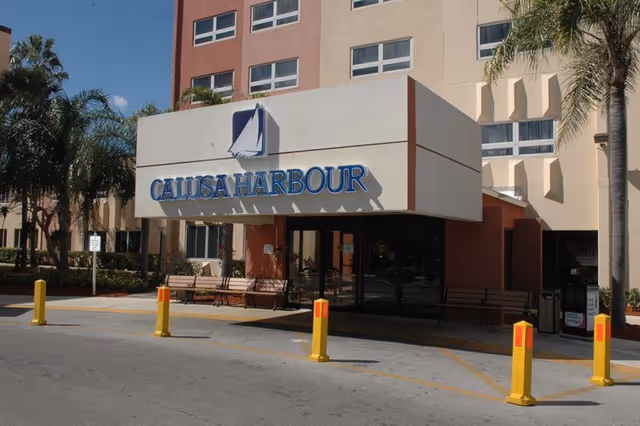 Entrance of Calusa Harbour facility with a large sign displaying the name and a sailboat logo above the doorway. The building is multi-story with several windows, palm trees on either side, benches near the entrance, and yellow bollards in front of the driveway.