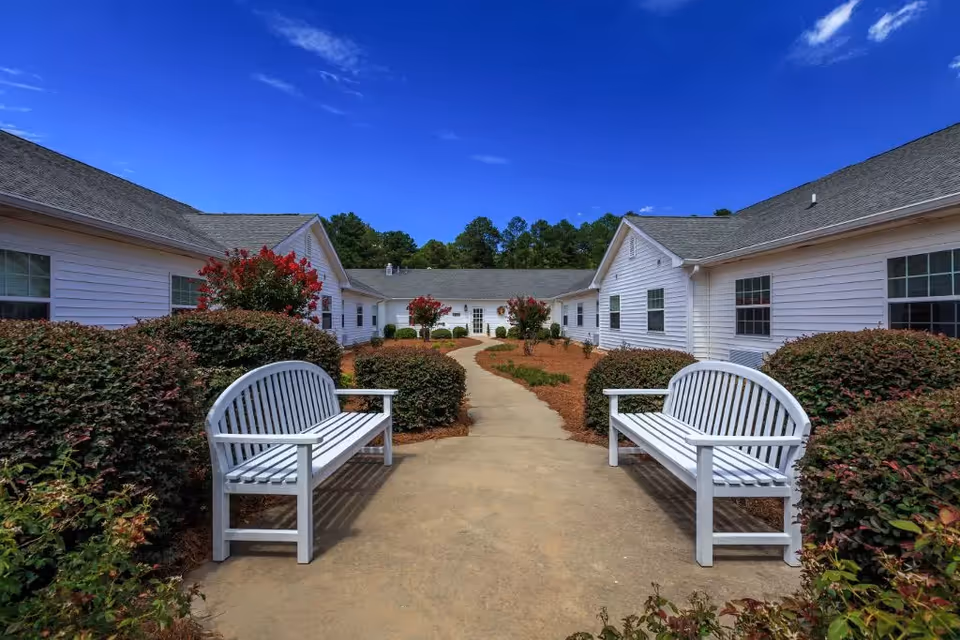 Outdoor courtyard area of Merryvale Assisted Living featuring a paved walkway flanked by two white benches and surrounded by bushes and flowering plants, with white buildings on either side under a clear blue sky.