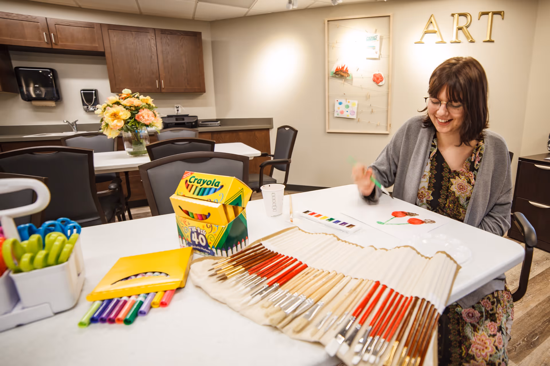 A woman with glasses and a floral dress sits at a table in an art room, smiling as she paints with watercolors. The table is covered with various art supplies including paintbrushes, colored markers, and a box of Crayola colored pencils. Behind her on the wall is a display board with artwork and the word 'ART' in large gold letters. The room has wooden cabinets, a sink, and a vase of flowers on another table in the background.