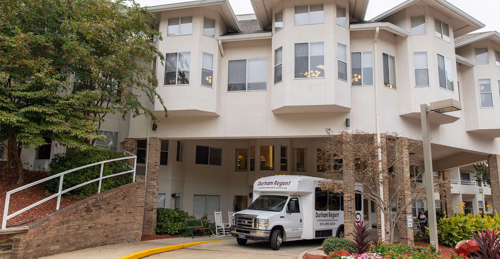 Front entrance of a multi-story senior living building with a covered drop-off, landscaping, and a white Durham Regent shuttle van parked under the canopy.