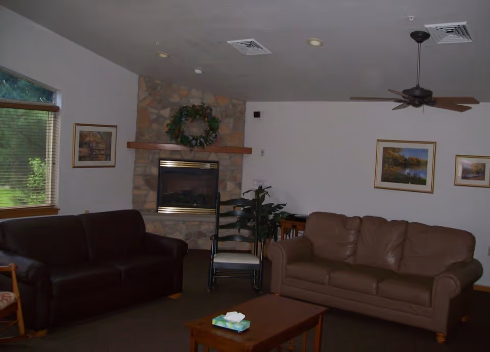 A cozy living room with two sofas, one dark brown and one light brown, a wooden coffee table with a tissue box, a stone fireplace decorated with a green wreath, a ceiling fan, framed landscape paintings on the wall, and a window with blinds showing greenery outside.