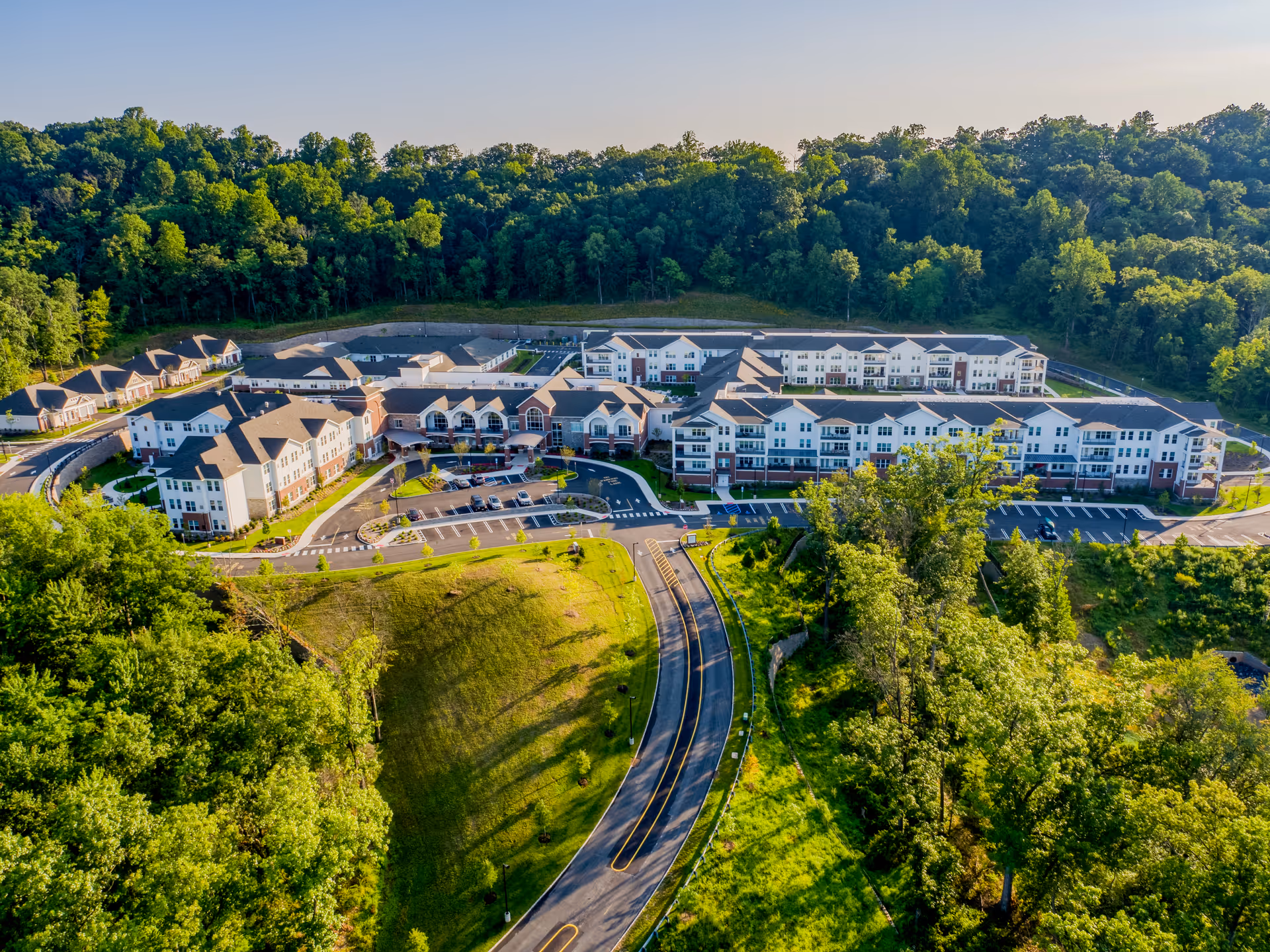 Aerial view of The Delaney of Bridgewater senior living facility surrounded by lush green trees and landscaping, showing multiple connected buildings with parking lots and a curved road leading to the entrance.