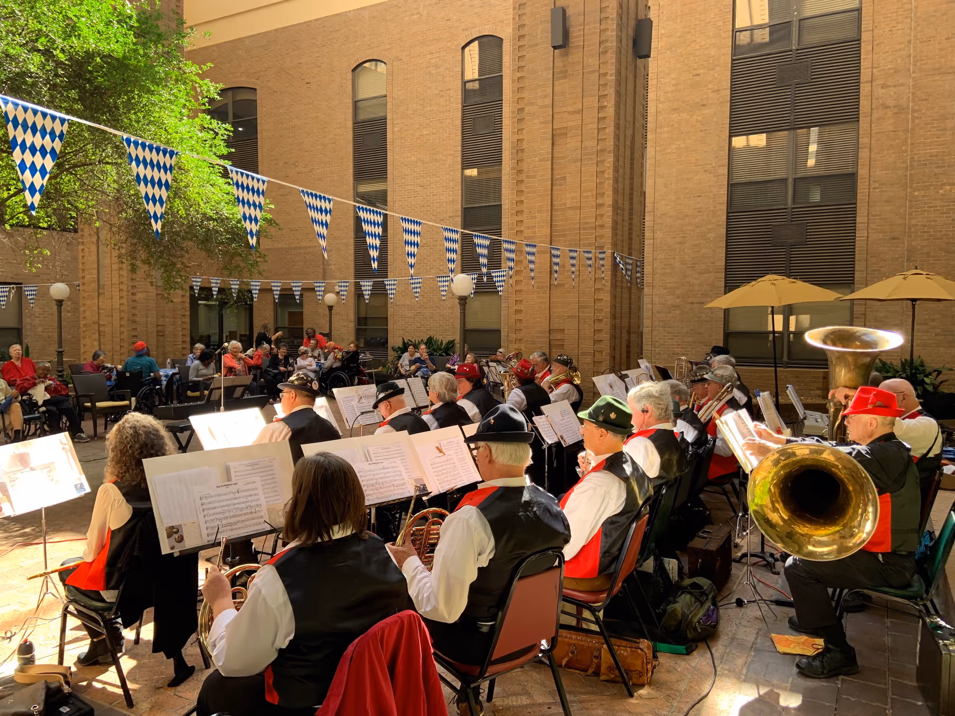 An outdoor courtyard at Walnut Place where a band of elderly musicians dressed in traditional attire is performing. The musicians are seated in rows with music stands in front of them, playing various brass and woodwind instruments. Blue and white checkered pennant banners are strung overhead, and several elderly audience members are seated around the courtyard watching the performance. The courtyard is surrounded by tall brick walls with windows, and there are a few large umbrellas providing shade.