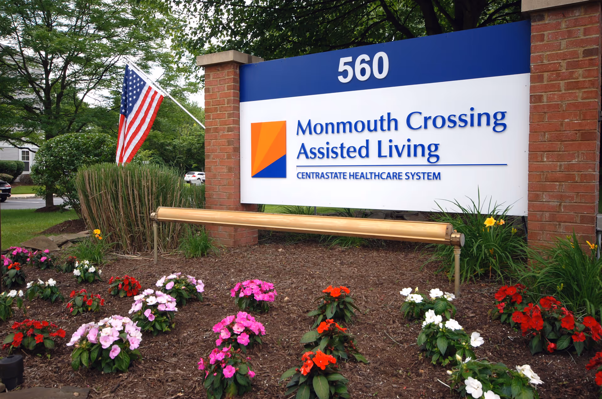 Outdoor sign for Monmouth Crossing Assisted Living, part of CentraState Healthcare System, with the address number 560 displayed above. The sign is set between two brick pillars with a garden bed of colorful flowers in front and an American flag on a pole to the left.