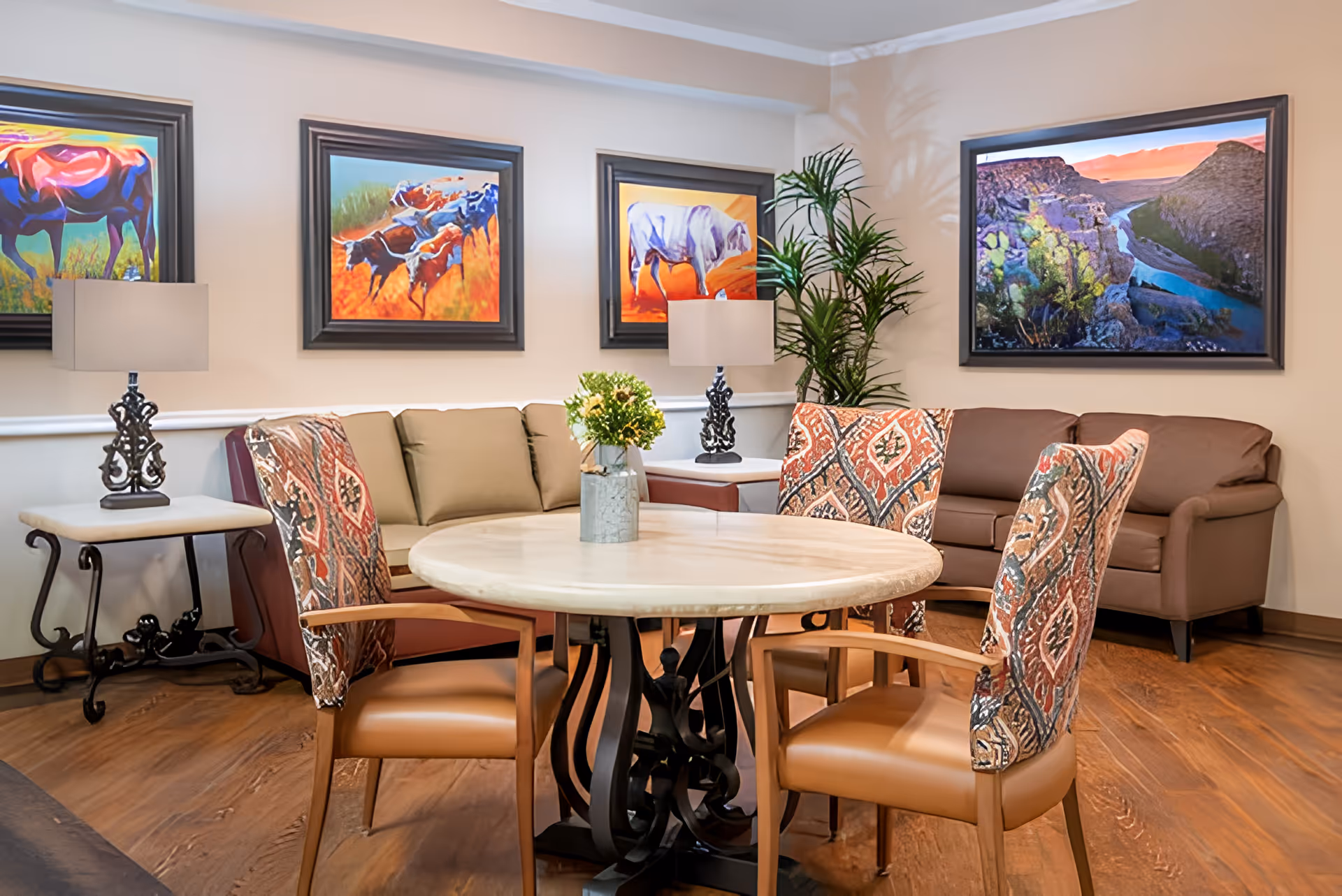 A bright communal living room with a round table and patterned chairs, sofas, lamps, potted plant, and colorful framed artwork on the walls.