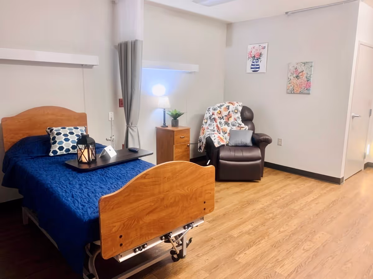 A tidy nursing facility patient room with a single hospital bed dressed in blue, a bedside table and lamp, and a recliner chair draped with a floral blanket.