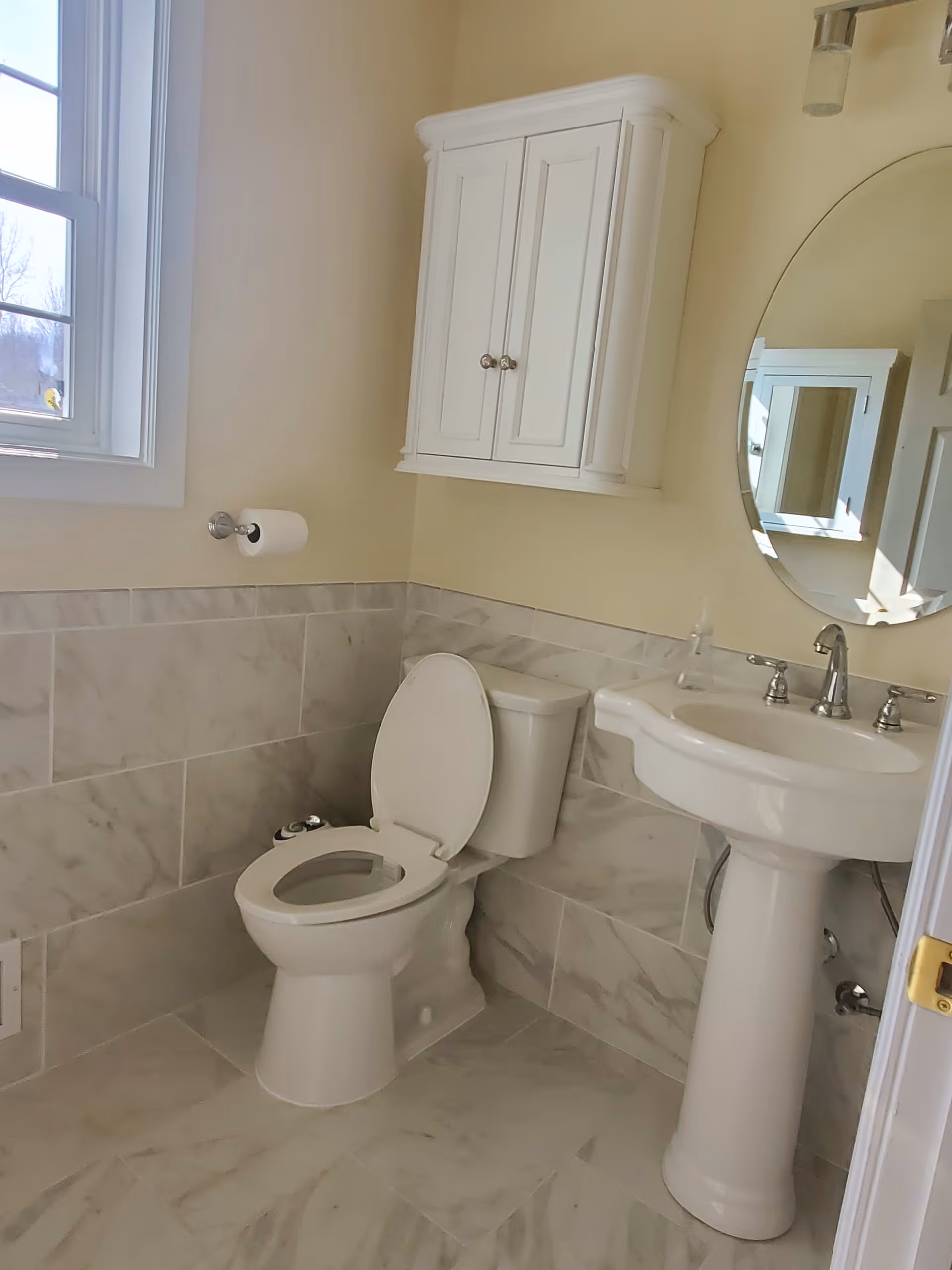 A clean bathroom with a white toilet with the lid open, a white pedestal sink with silver faucets, a round mirror above the sink, a white wall-mounted cabinet, and a window letting in natural light. The walls and floor are tiled with light-colored marble tiles.