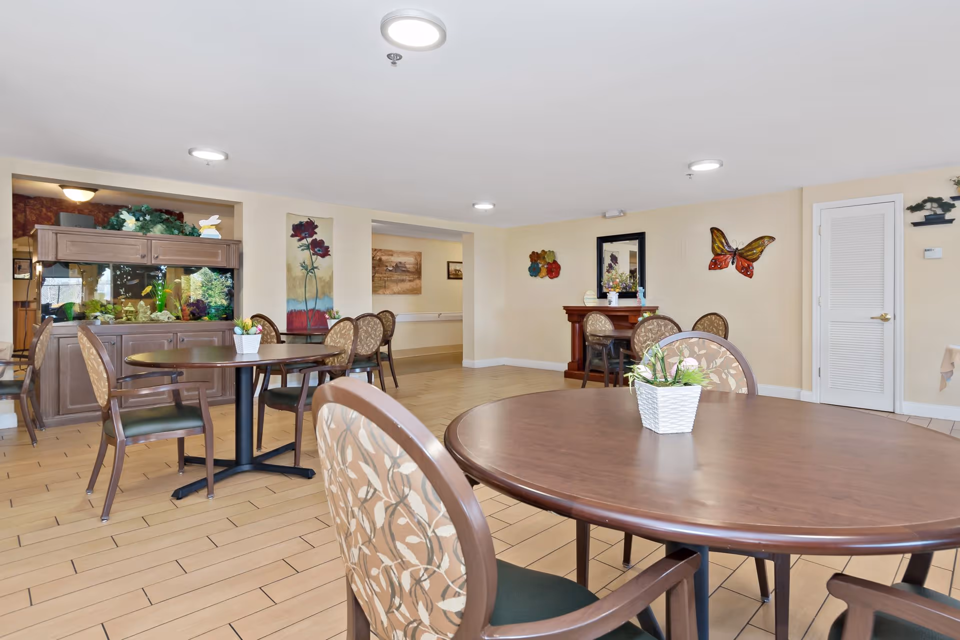 Bright communal dining room with round wooden tables, patterned chairs, potted plants and wall decorations.