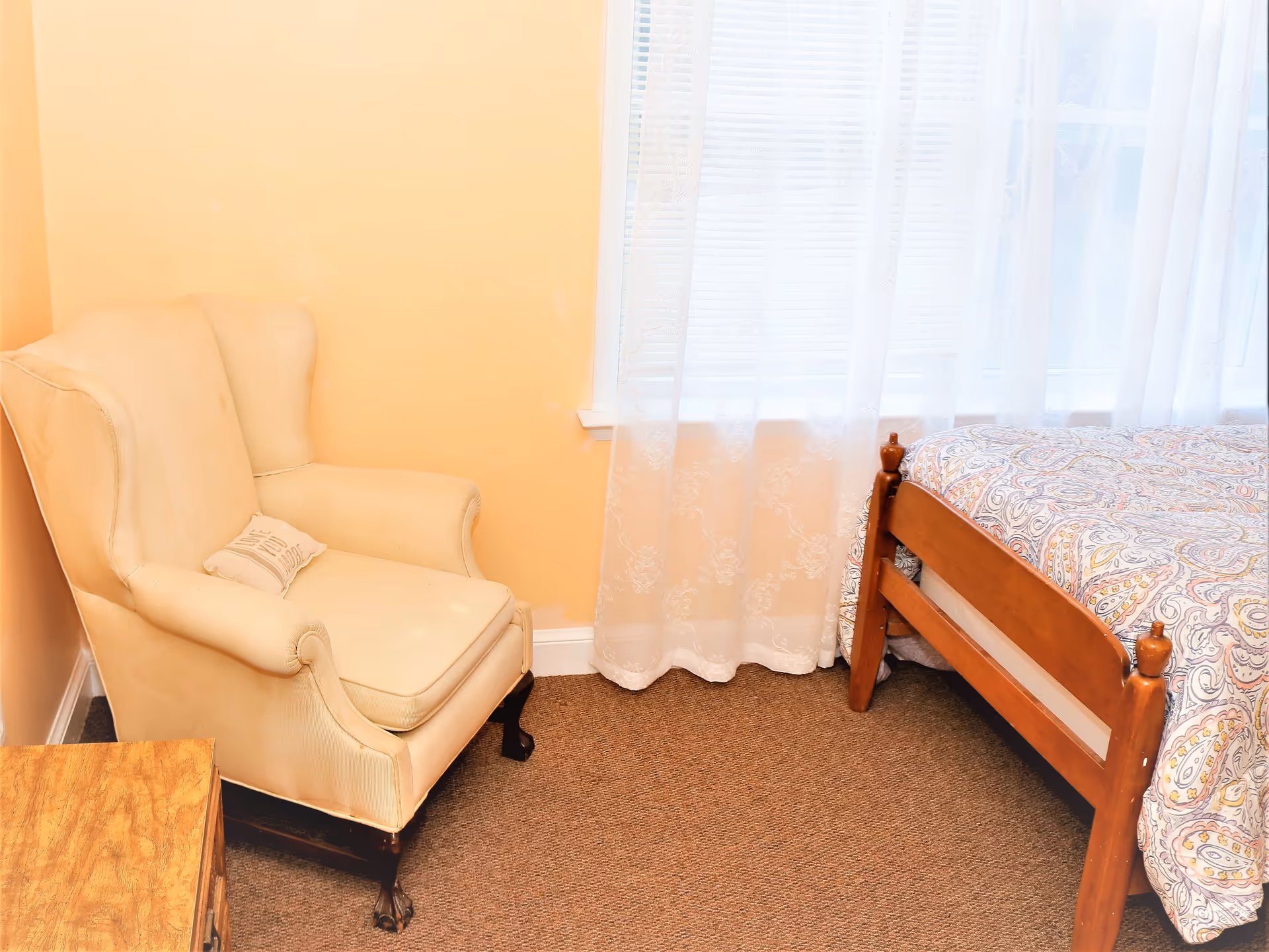 A cozy corner of a bedroom featuring a beige upholstered armchair with a small pillow, a wooden bed frame with a patterned bedspread, and a window with sheer white curtains allowing natural light to enter.