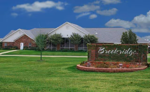 Front lawn and sign reading 'Brookridge' with a single-story brick and white retirement community building in the background under a blue sky.