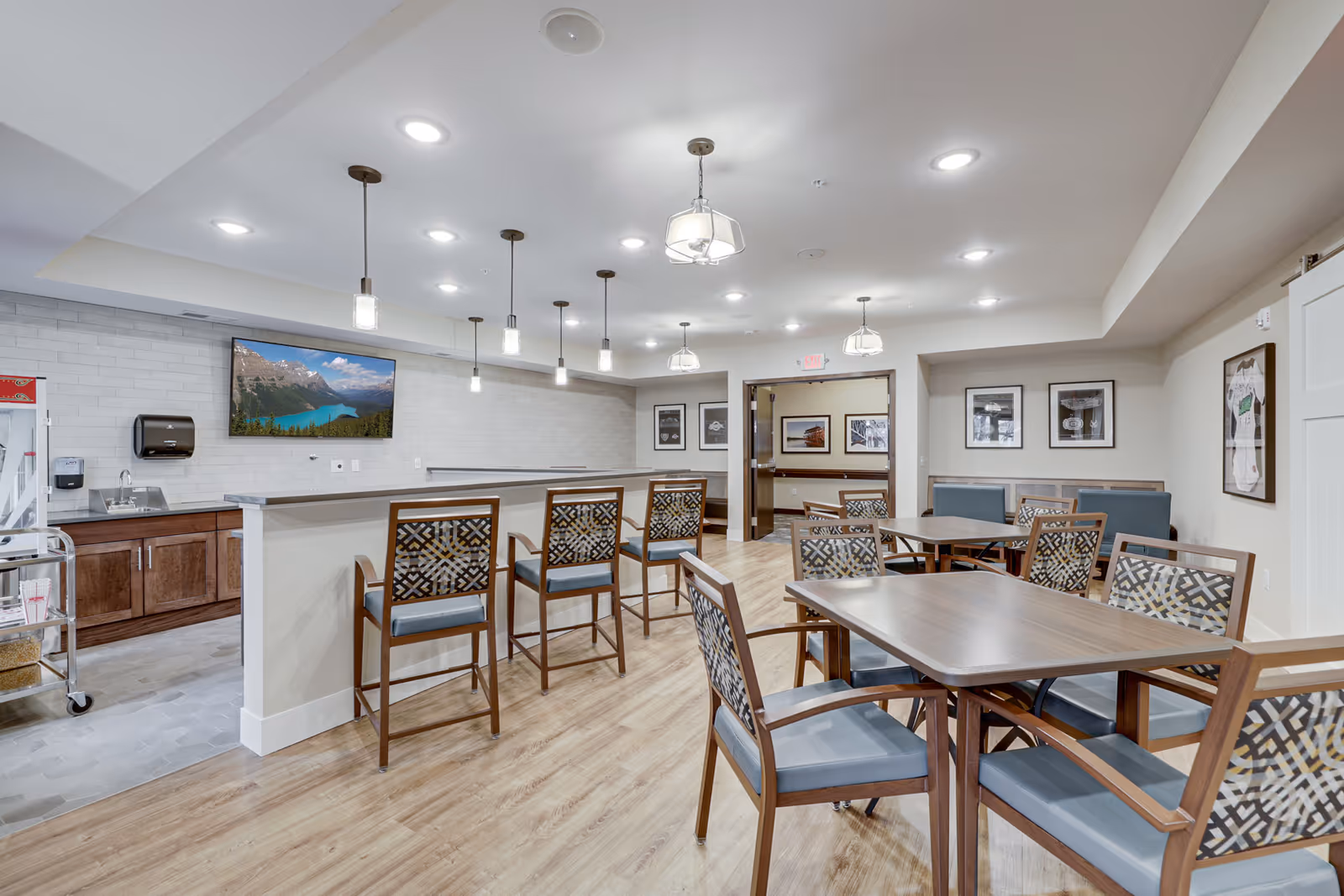 Well-lit communal dining area with tables and patterned chairs, a high counter with bar stools, pendant lights, and a wall-mounted TV.