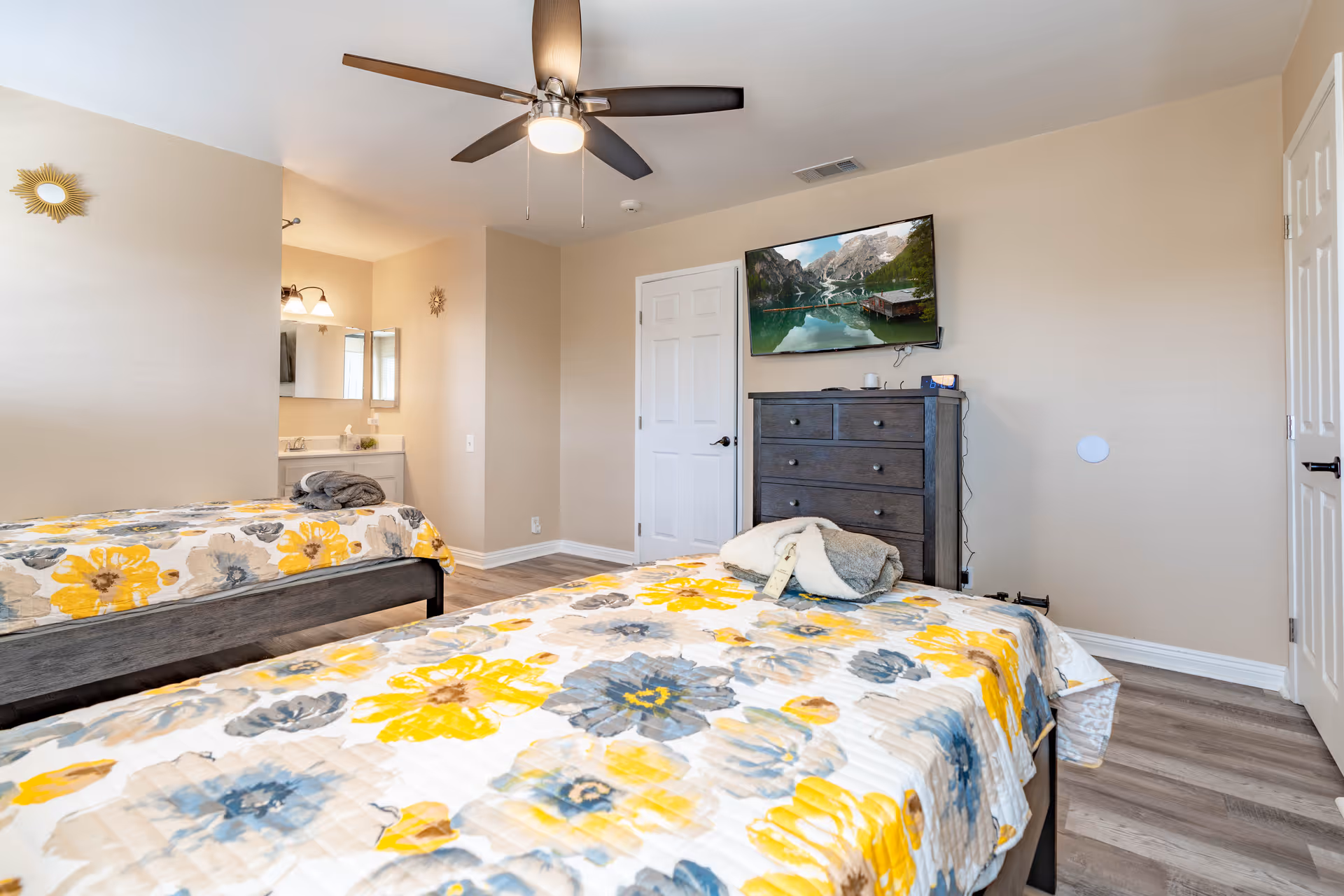 A bright bedroom with two twin beds covered in floral bedspreads featuring yellow, gray, and blue flowers. The room has light beige walls, a ceiling fan with lights, a dark wooden dresser with a flat-screen TV mounted above it displaying a scenic mountain and lake view. There is a small vanity area with a mirror and light fixtures visible through an open doorway.