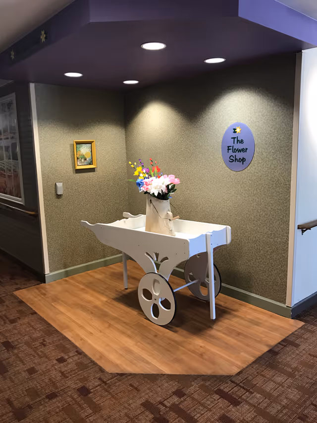 A white decorative cart with large wheels holding a vase of colorful artificial flowers is placed in a corner of a hallway with patterned wallpaper and wooden flooring. Above the cart, a purple oval sign reads 'The Flower Shop'. The ceiling has recessed lighting illuminating the area.