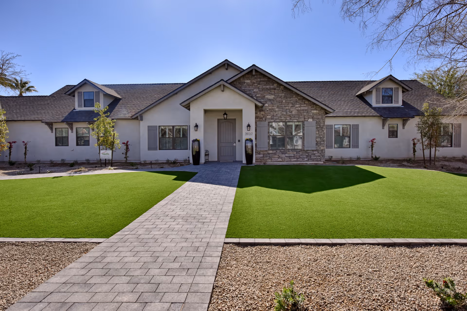Front exterior view of a single-story building with a stone and stucco facade, a central entrance with a covered porch, two large decorative vases on either side of the door, and a paved walkway leading up to the entrance. The building is surrounded by green artificial turf and gravel landscaping under a clear blue sky.