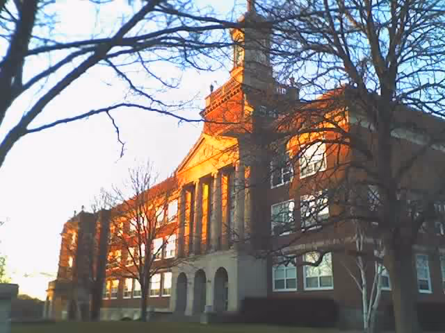 Front facade of a multi-story brick building with columns and arched entryways lit by warm sunlight, framed by leafless trees.