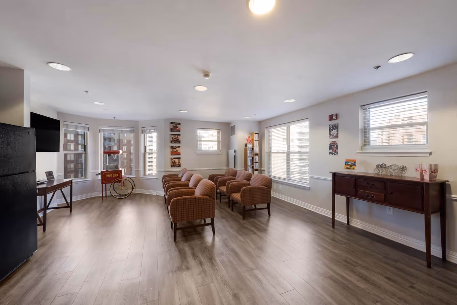 A bright and spacious common room with several cushioned chairs arranged in rows facing a wall-mounted TV. The room features large windows letting in natural light, a popcorn machine, a wooden sideboard with decorative items, and light wood flooring.