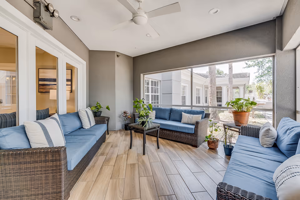 Bright screened sunroom with wicker sofas covered in blue cushions, a central coffee table and potted plants overlooking a courtyard.
