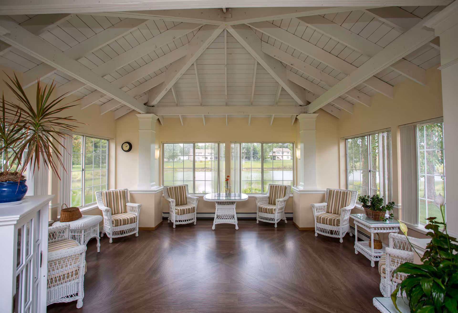 Bright sunroom-style common area with white wicker chairs around a central table and large windows overlooking a pond.
