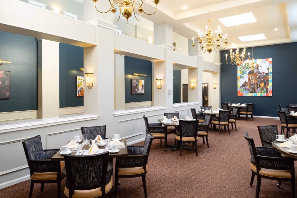 A spacious dining room with multiple tables set for meals, each with chairs, white napkins, cups, and utensils. The room features dark blue accent walls, decorative wall sconces, chandeliers hanging from the ceiling, and colorful artwork on the far wall. The carpet is brown, and the overall ambiance is elegant and inviting.