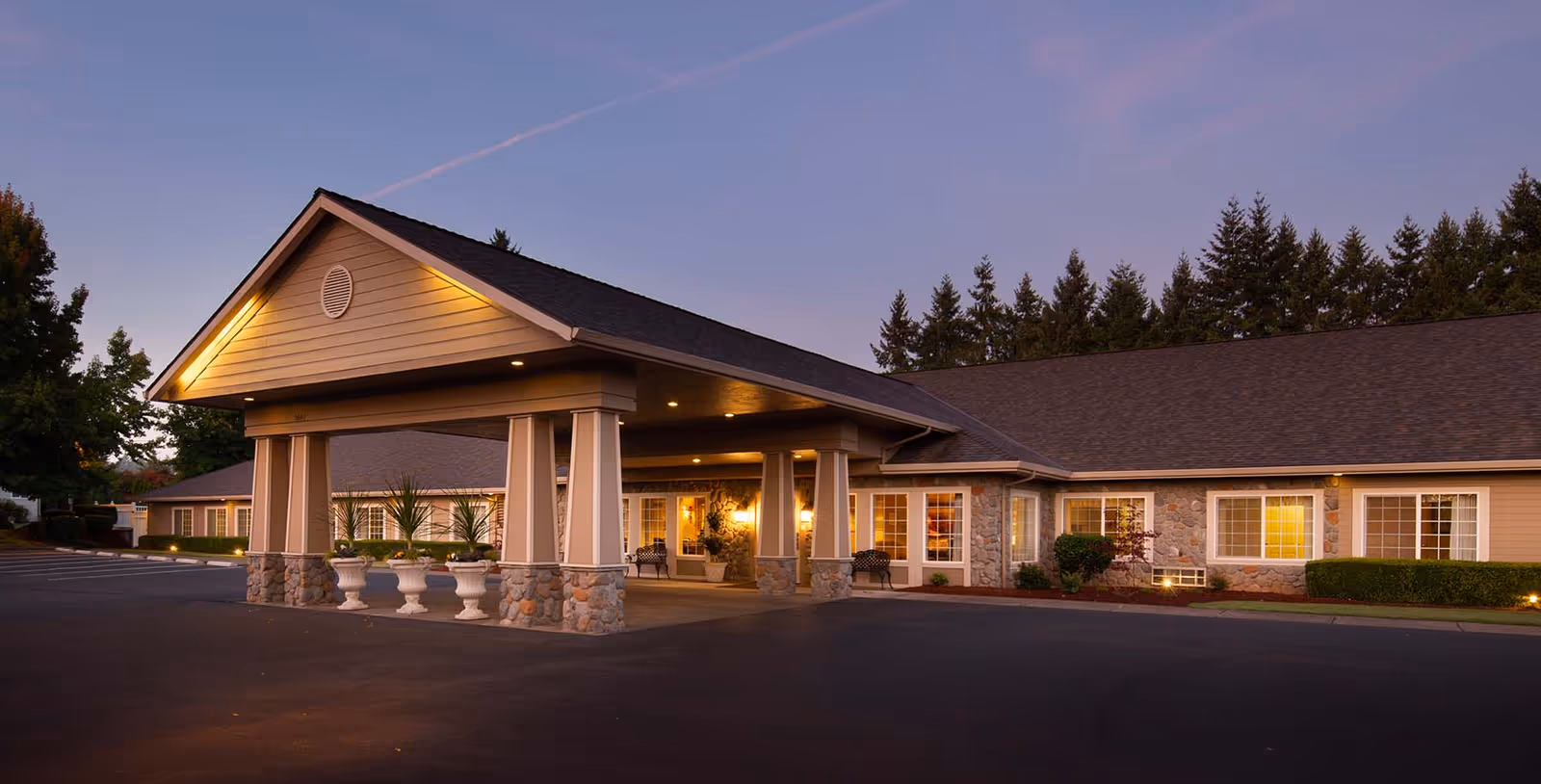 Exterior view of Cedar Creek of Bloomington Memory Care facility at dusk, showing the entrance with a covered driveway supported by stone and wooden pillars, illuminated by warm lights. The building has a stone and siding facade with multiple windows and is surrounded by trees and landscaping.