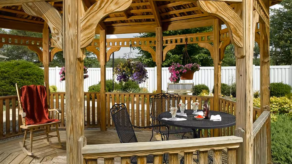 Wooden gazebo with a round black metal table and four matching chairs. On the table are three ice cream sundaes, a bottle of syrup, a small bowl of cherries, and a napkin. A wooden rocking chair with a red blanket draped over it is positioned inside the gazebo. Hanging flower pots with pink and purple flowers are suspended from the gazebo roof. The background shows a white fence, green bushes, and trees.