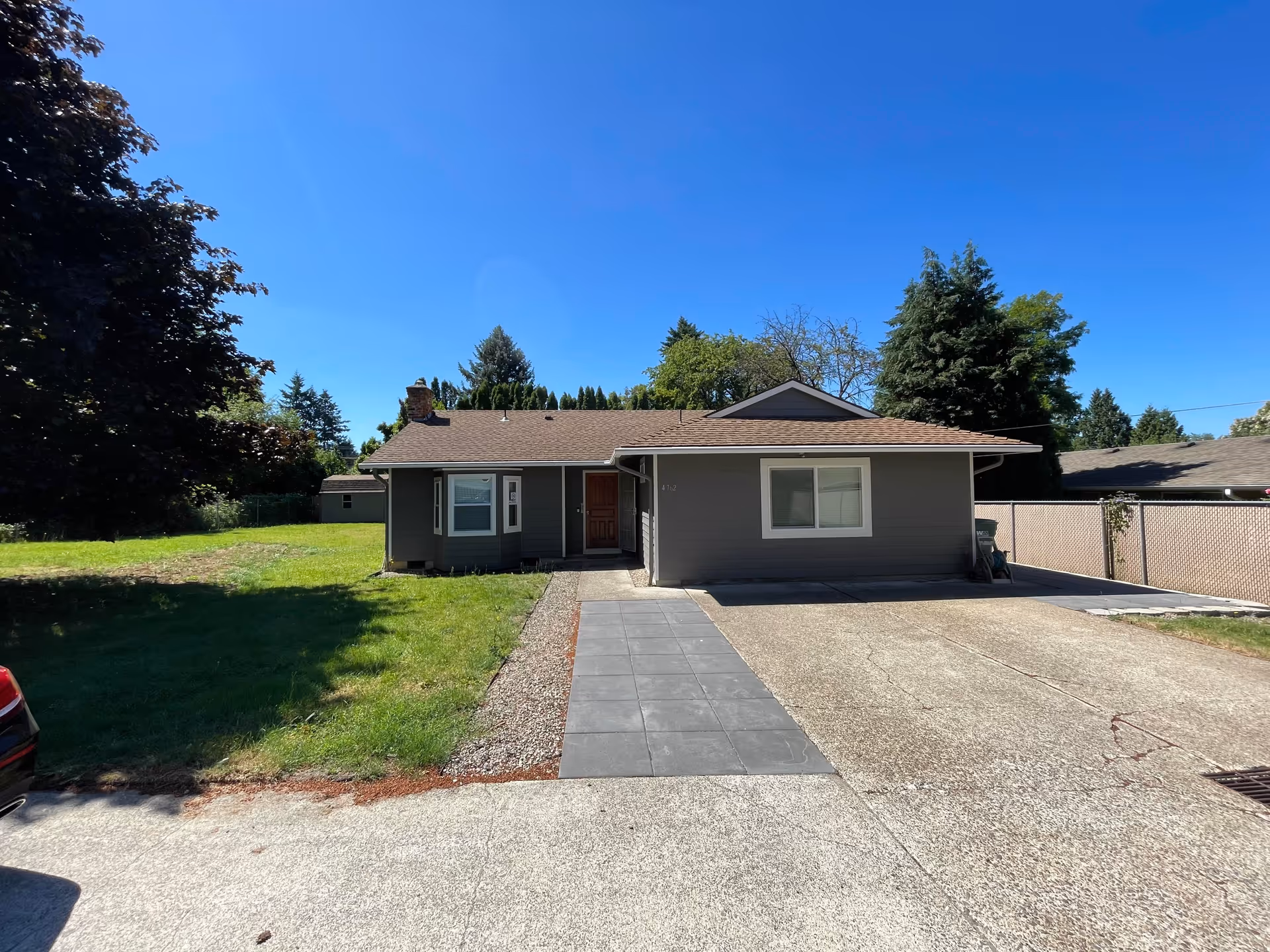 Single-story gray house with a brown roof, a front door, and two windows. There is a paved driveway and a tiled walkway leading to the entrance. The house is surrounded by green grass, trees, and a clear blue sky.