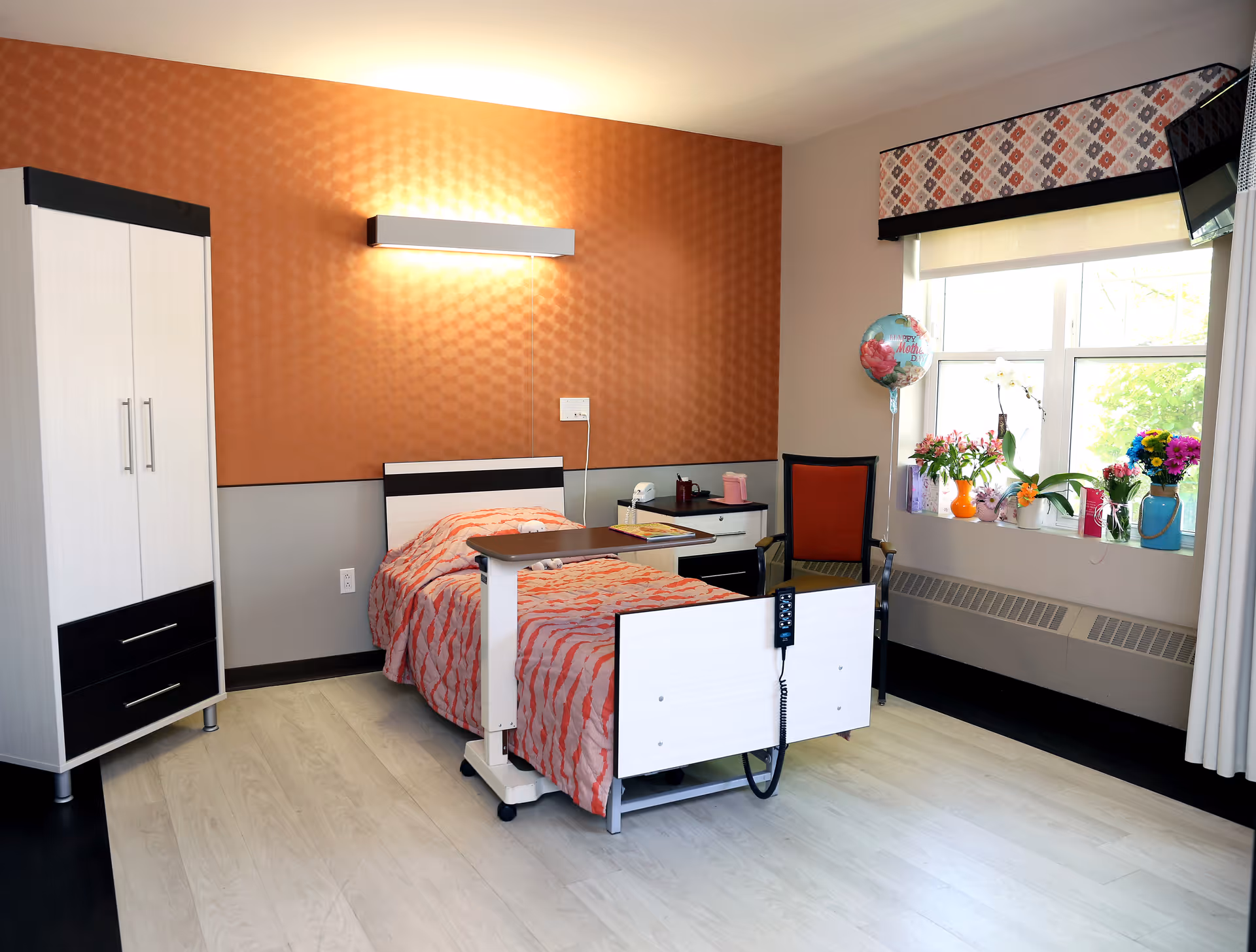 A bright and clean senior living bedroom with a single bed covered in a red and white patterned blanket. The room features a white and black wardrobe, a bedside table with items on it, a red chair, and a window with floral decorations and a Happy Mother's Day balloon. The walls are painted with an orange textured upper half and a gray lower half, and there is a modern light fixture above the bed.
