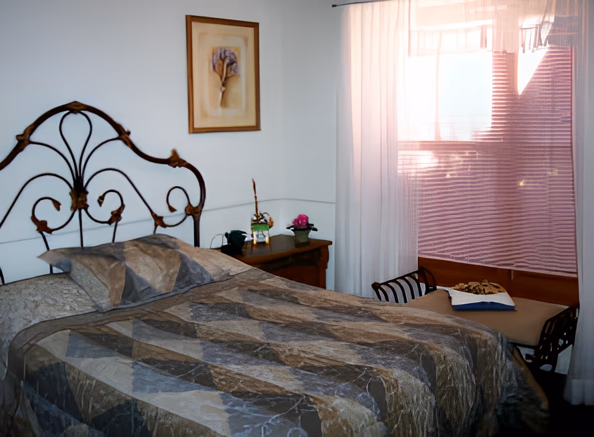 A bedroom with a decorative metal headboard, a bed covered with a patterned quilt, a wooden nightstand with small plants and a framed picture on the wall. There is a window with sheer curtains allowing light to enter the room.