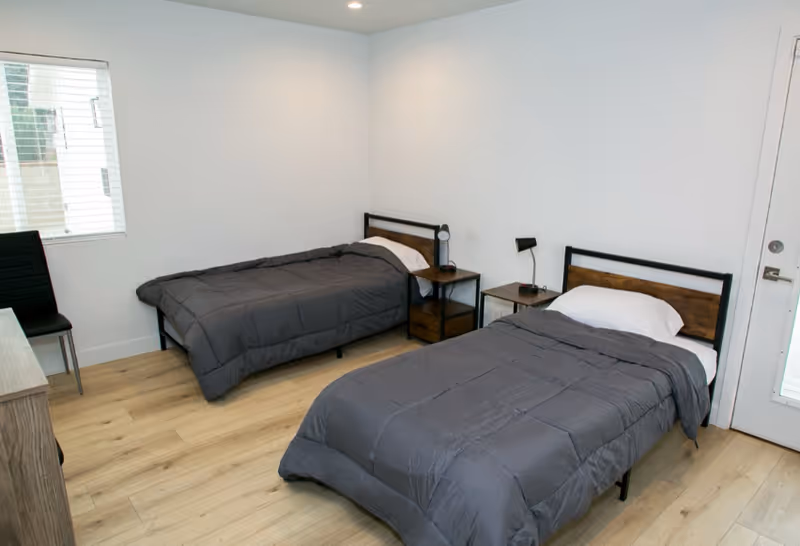 Two single beds with gray quilts and matching nightstands in a bright bedroom.