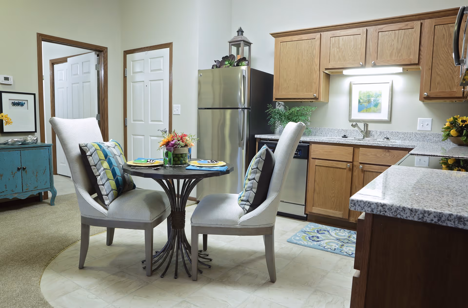 A cozy kitchen and dining area in a retirement community apartment. The kitchen features wooden cabinets, a stainless steel refrigerator, dishwasher, and granite countertops. A small round dining table with two cushioned chairs, each with a decorative pillow, is set with plates and napkins. There is a blue sideboard against the wall and a framed picture above the sink. The floor transitions from carpet in the living area to tile in the kitchen.