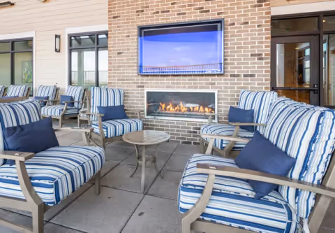 Outdoor seating area with striped cushioned chairs and a small round table arranged around a modern rectangular gas fireplace set in a brick wall. Above the fireplace is a mounted flat-screen TV. The area is part of a covered patio with large windows and doors leading inside.