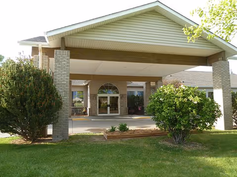 Front exterior view of Highland Park Assisted Living building with a covered entrance supported by brick pillars, surrounded by green bushes and a well-maintained lawn.