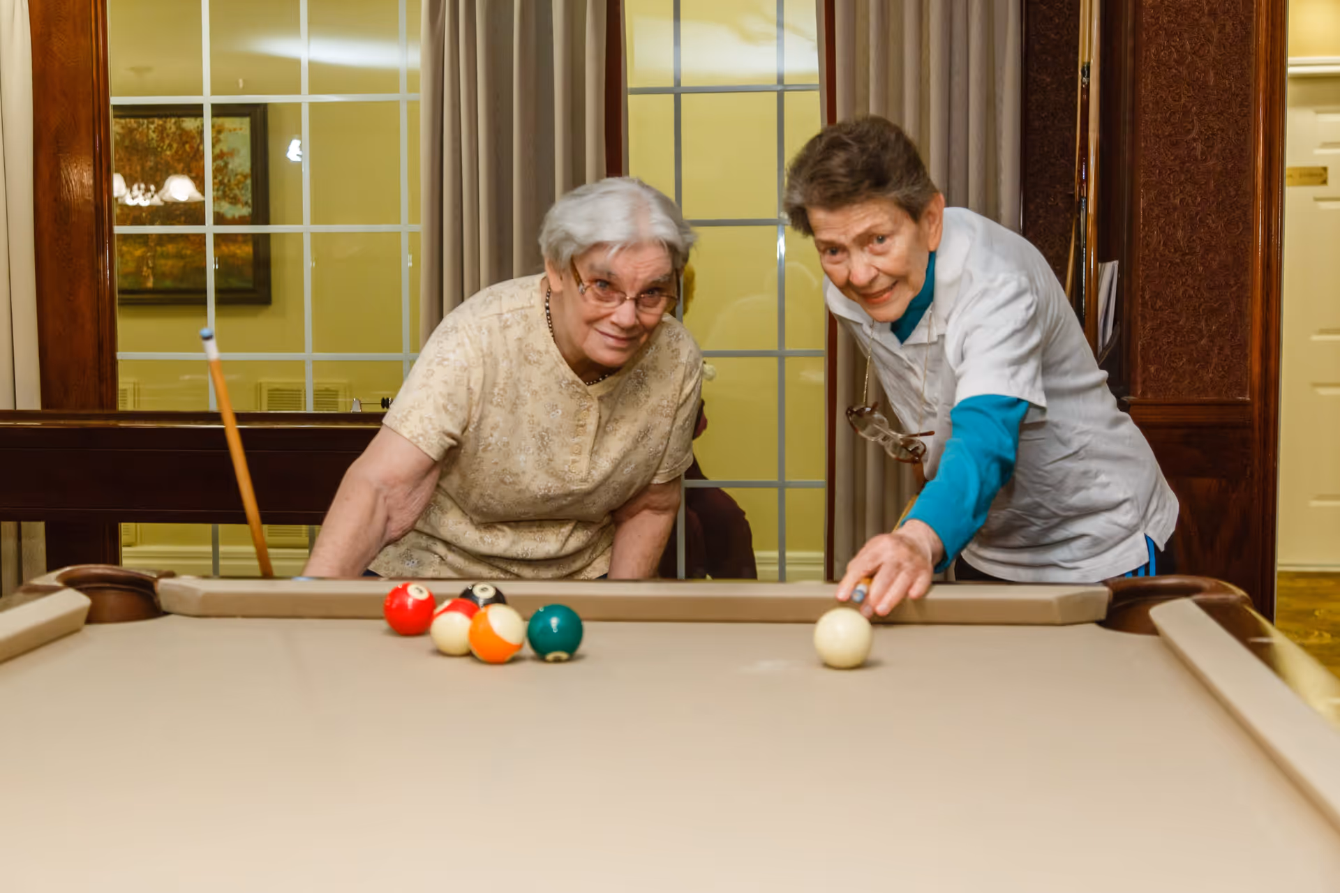 Two elderly women playing pool indoors, one preparing to strike the cue ball while the other watches and smiles. The room has large windows with curtains and warm lighting.