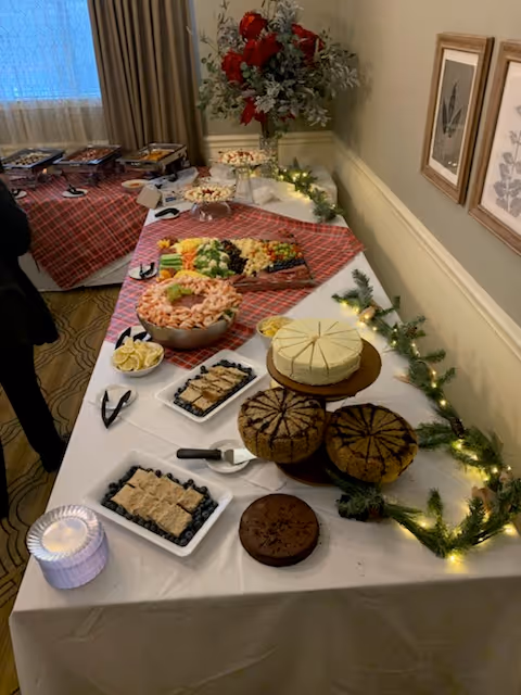 A buffet table set up with various foods including a vegetable and cheese platter, a bowl of shrimp with lemon wedges, several cakes on a tiered stand, and trays of other appetizers. The table is decorated with greenery and string lights, and there are framed botanical prints on the wall behind it.