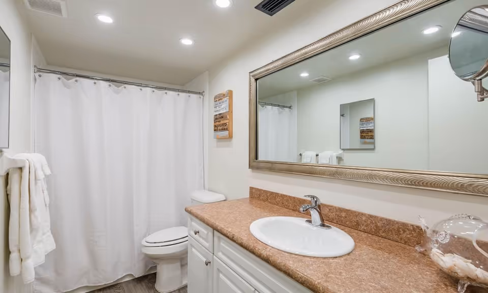 A clean and well-lit bathroom with a white shower curtain, a toilet, a countertop with a sink, a large framed mirror above the sink, and white towels hanging on a rack.