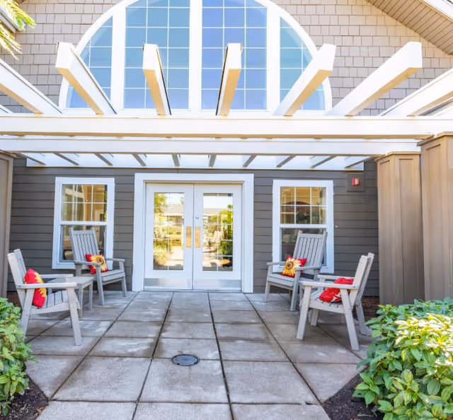 Front entrance patio with a pergola, double glass doors, and four wooden chairs with colorful cushions.