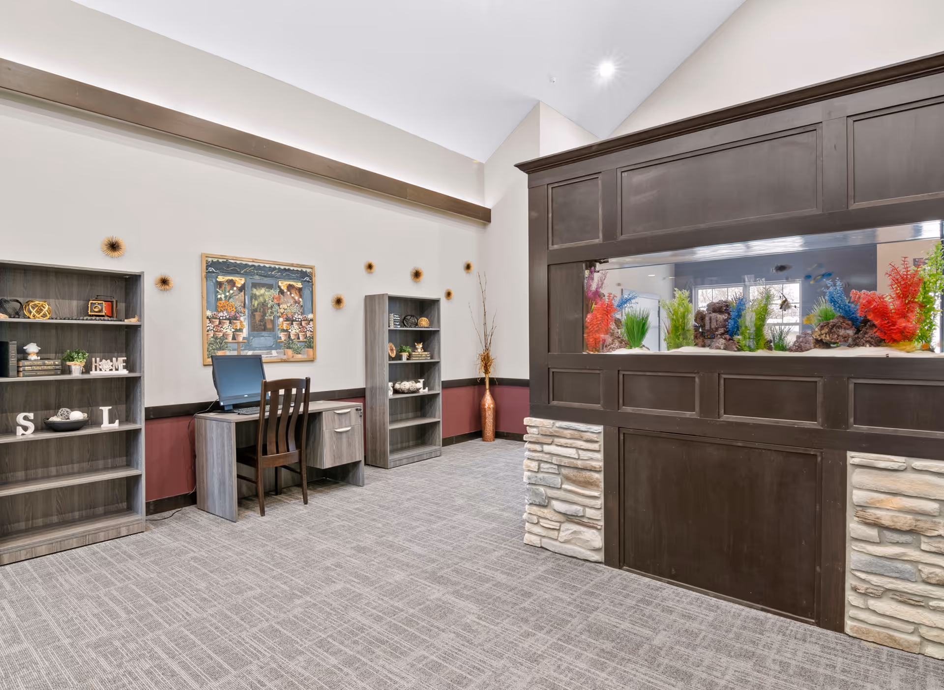 A well-lit interior room with a large built-in aquarium on the right side, featuring colorful aquatic plants and rocks. On the left side, there are two gray wooden bookshelves with decorative items and a small desk with a computer and chair. The walls are light-colored with a maroon accent below the chair rail, and a framed painting hangs above the desk. The floor is carpeted in a neutral tone.